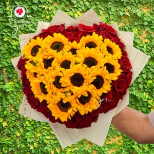 Arreglo en forma de corazón con girasoles y rosas rojas sobre un fondo natural verde.