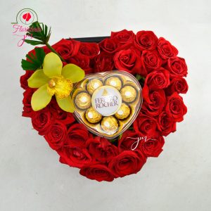 Arreglo floral en forma de corazón con rosas rojas, chocolates Ferrero Rocher y una orquídea amarilla.