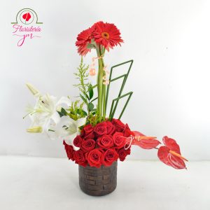 Arreglo floral vertical en base rústica con rosas rojas, lirios blancos y gerberas.