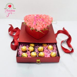 Caja transparente con un arreglo de rosas rosadas y una tapa decorativa de Feliz Cumpleaños, acompañada de chocolates Ferrero Rocher