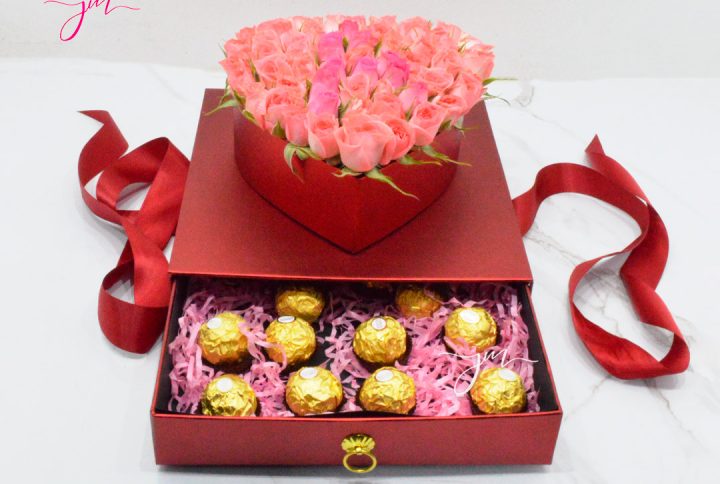 Caja transparente con un arreglo de rosas rosadas y una tapa decorativa de Feliz Cumpleaños, acompañada de chocolates Ferrero Rocher