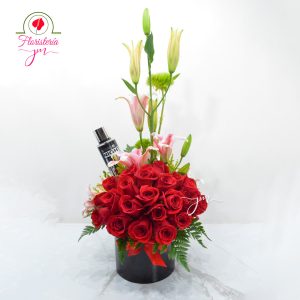 Arreglo floral con rosas rojas, lirios rosados y detalles verdes, acompañado de un perfume en base negra.