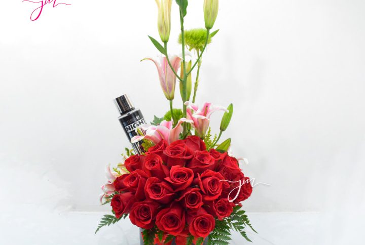 Arreglo floral con rosas rojas, lirios rosados y detalles verdes, acompañado de un perfume en base negra.