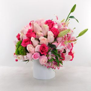 Arreglo floral elegante con rosas rosadas, lirios y detalles verdes en base blanca.