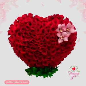 Arreglo floral en forma de corazón con rosas rojas sobre una base blanca decorada con detalles navideños y un lazo rojo.